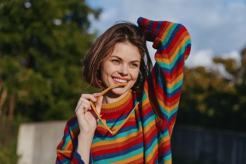 Woman with a bright smile, portrait in a colorful sweater outdoors holding sunglasses and posing casually, natural light, short hair, playful cozy autumn fashion and joyful expression.