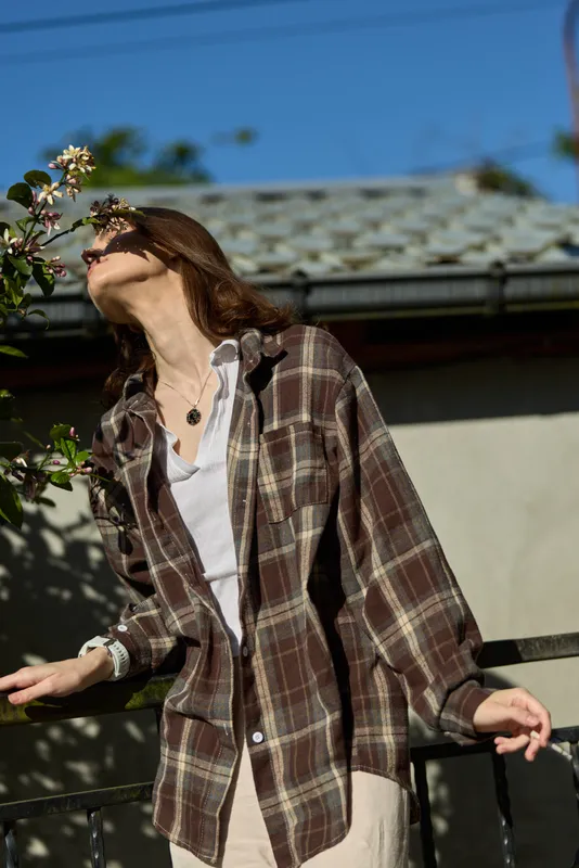 Casual woman in a plaid shirt enjoys a sunny moment tilting her head toward a flower as breeze moves her hair, capturing a relaxed outdoor lifestyle scene