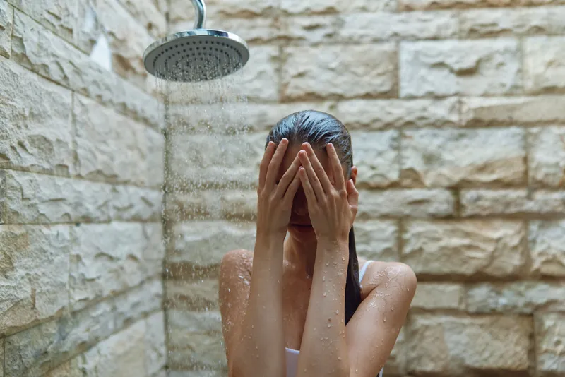woman taking shower outdoors with stone wall background, wet hair, natural light, summer, self-care, water droplets, refreshing moment
