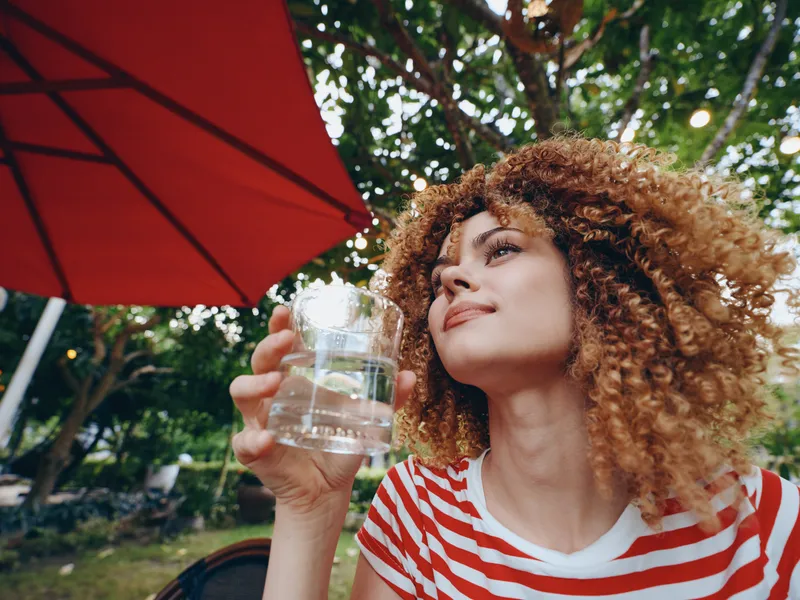 Portrait of a young woman with curly hair enjoying a glass of water outdoors, sitting under a red umbrella, sunlight filtering through trees and creating a warm, relaxed summer mood.