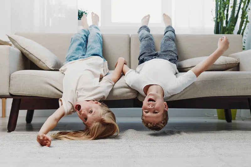Two happy children lie upside down on a sofa holding hands, laughing joyfully in a bright living room. Concept of friendship, childhood fun, carefree moments, and indoor playtime.