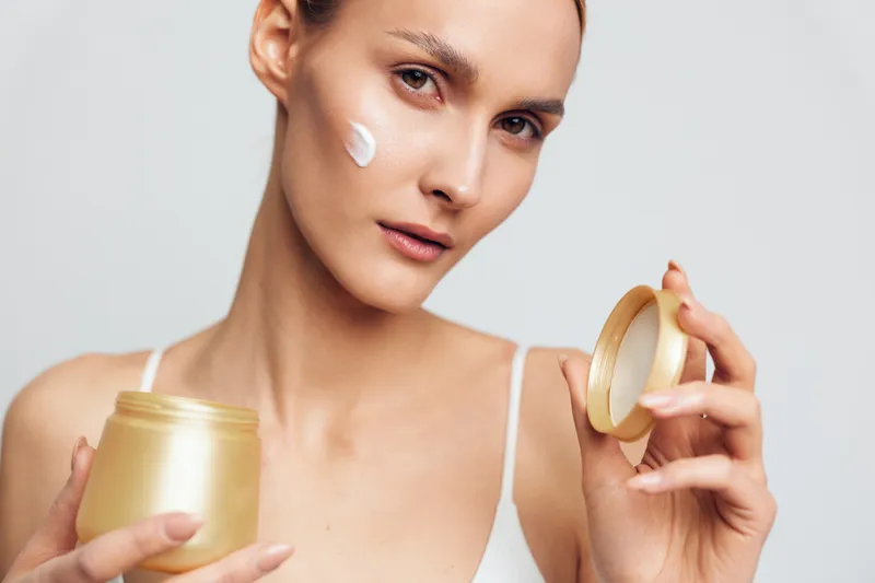 Young woman applying face cream with soft texture, clear skin, natural beauty, skincare routine, neutral background, close up, healthy lifestyle concept