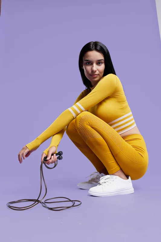 Sport fitness woman in vibrant yellow activewear squats holding a jump rope, showcasing strength, focus, and athletic lifestyle against a purple background.