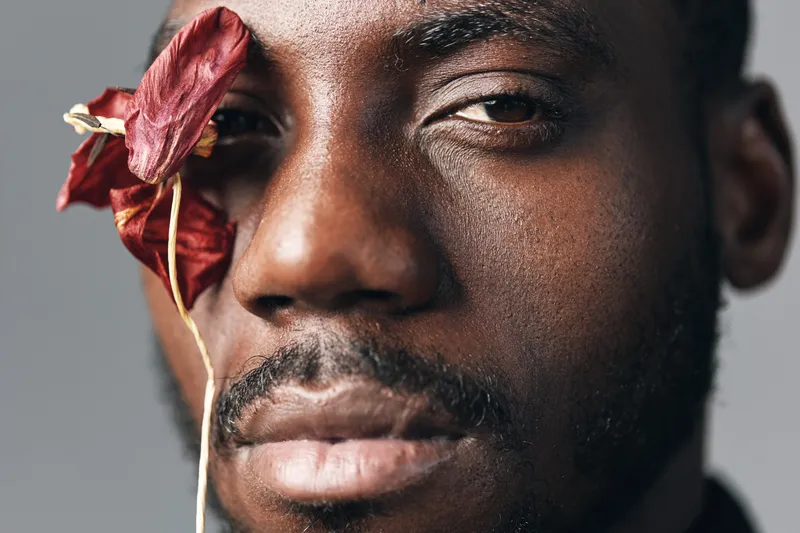 Closeup portrait of black African man with dark skin and beard holding dried red flower near eye in creative visual designer style on gray background for artistic expression and beauty.
