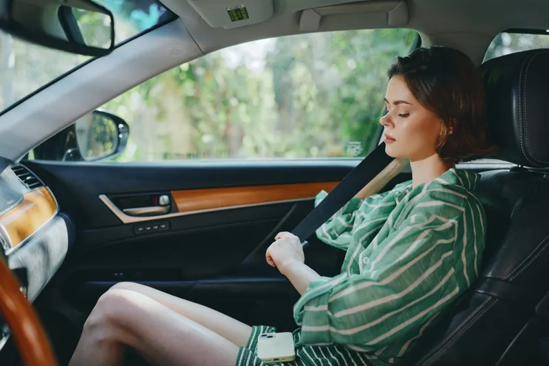 A woman sits in a car wearing a seat belt, gazing calmly ahead. She wears a green striped dress that contrasts with the dark leather interior.