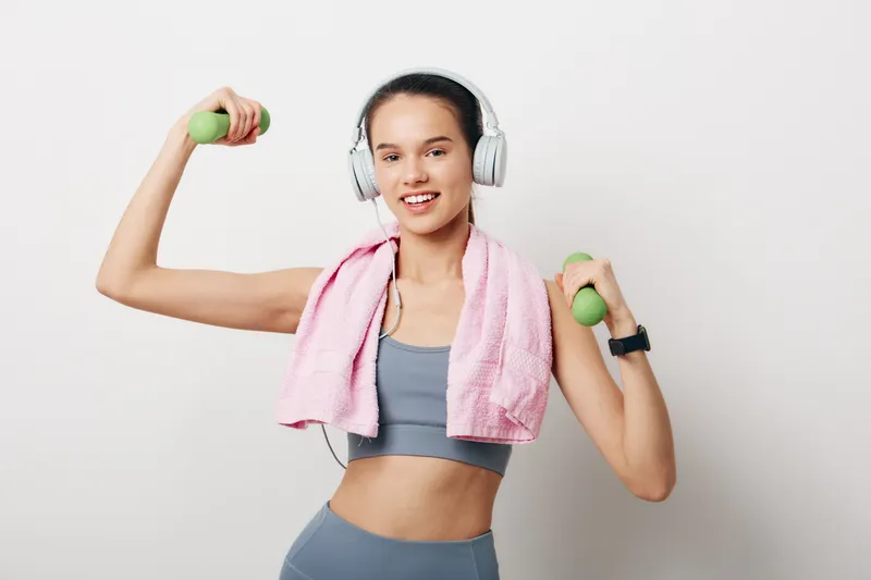 Woman fitness, sport: young woman fitness portrait holding green dumbbells, wearing gray sports bra and leggings, pink towel over shoulders, white headphones, smiling on white background.