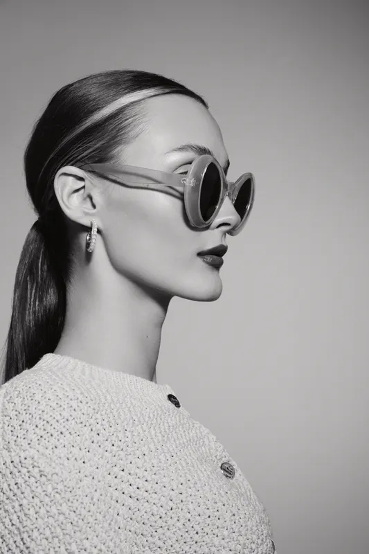 Profile of a caucasian woman with light skin wearing large round sunglasses and a knitted top. Stylish female model poses in studio against plain background.