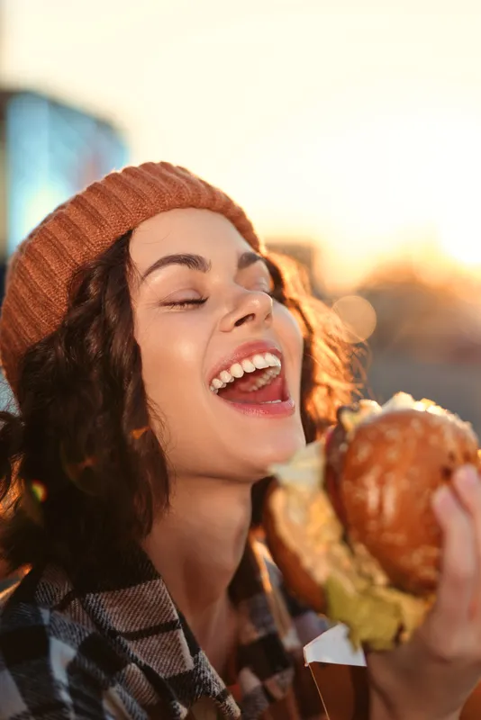 Woman holding burger outdoors with joyful smile and knit hat, candid lifestyle moment in golden hour glow; authenticity and emotional storytelling capture mindful living atmosphere.