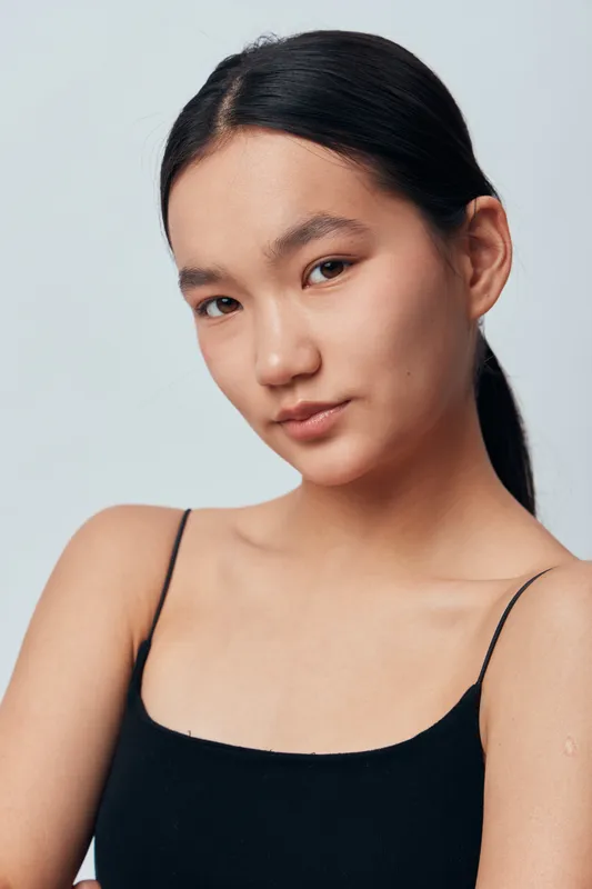 Young woman with natural skin tone and black hair tied back wearing black sleeveless top posing with calm expression in studio light on plain background. Beauty, health and fashion portrait.