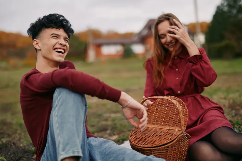 Two friends share a candid outdoor moment, radiating authenticity and genuine joy as autumn light fills the park and a wicker basket sits nearby.