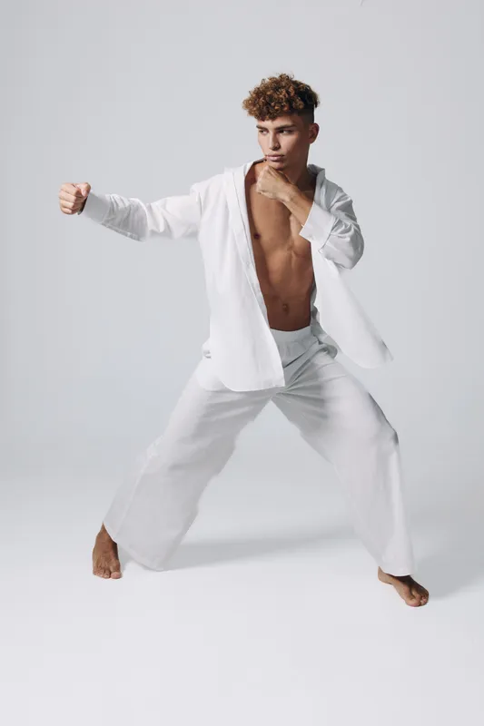 Young man practicing martial arts in white loose clothing, standing in fighting stance with focused expression on plain background. Fitness, training and discipline concept.