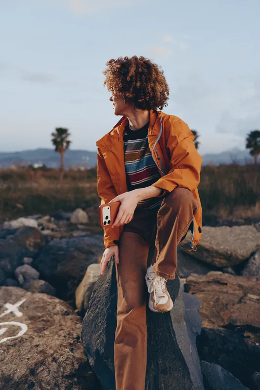 Woman lifestyle, outdoor walking on rocks, curly hair and orange jacket, holding a phone, relaxed confident expression, countryside landscape at sunset, natural daylight, lifestyle travel moment