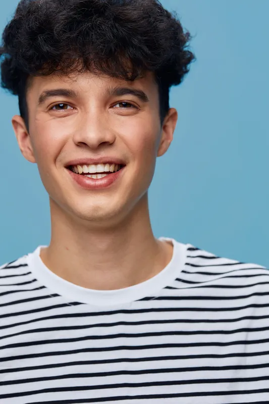 smiling man wearing striped shirt looks cheerful with curly hair against plain blue background, youthful and positive expression, casual style portrait
