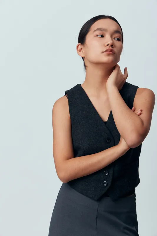 Young woman with long dark hair and sleeveless vest posing thoughtful and confident on light background. Portrait of female model with crossed arms and casual expression in studio.