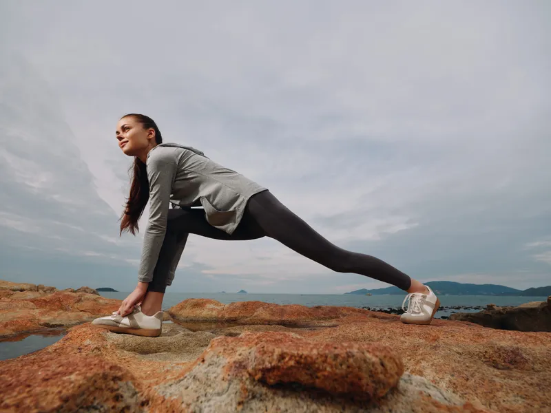 Young woman stretching on rocky terrain by the sea, dressed in activewear for fitness, showcasing a healthy lifestyle and determination during outdoor exercise