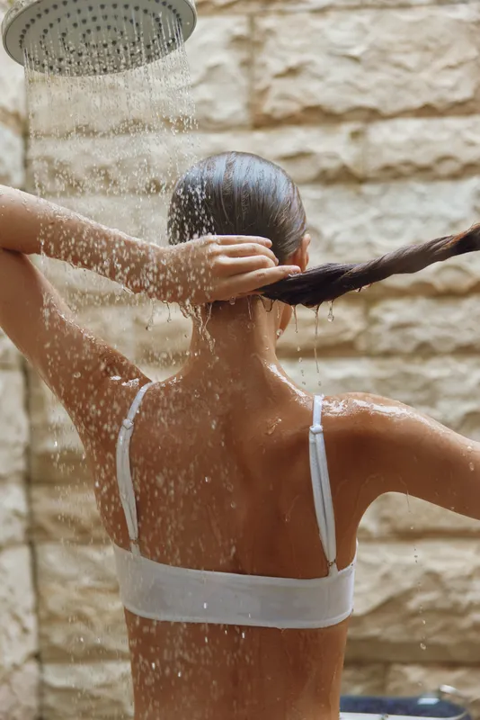 woman taking shower outdoors with flowing water, wet hair, stone wall background, natural light, refreshing moment, summer wellness.