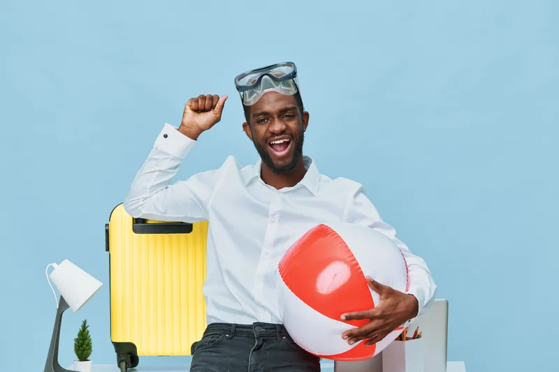 Enthusiastic young smiling man with dark skin holding a beach ball and wearing swim goggles, standing in front of travel accessories such as a yellow suitcase and a lamp, on bright pastel blue backgro