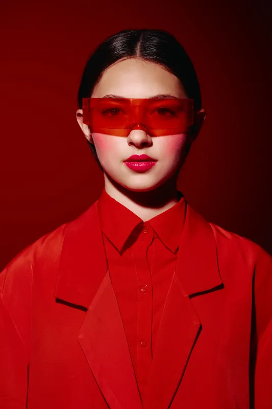Young woman wearing red tinted sunglasses and an oversized red jacket poses against a red background with bold makeup. Fashion concept with modern beauty and creative style.