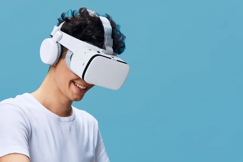 young man wearing virtual reality headset and white headphones smiling against blue background, immersive technology, casual white t-shirt, digital experience