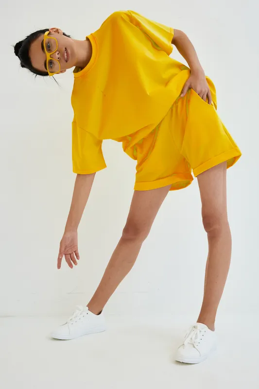 young woman in yellow oversized t-shirt and shorts poses in minimal white studio with casual white sneakers and yellow glasses, showcasing modern sporty street style outfit