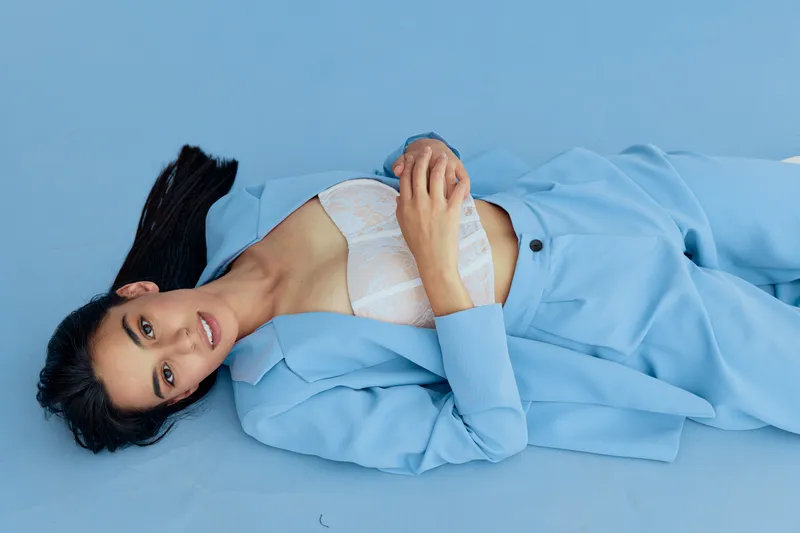 young woman lying on blue floor wearing pastel blue suit and white lace top, with long dark hair and calm expression in minimalistic studio setting