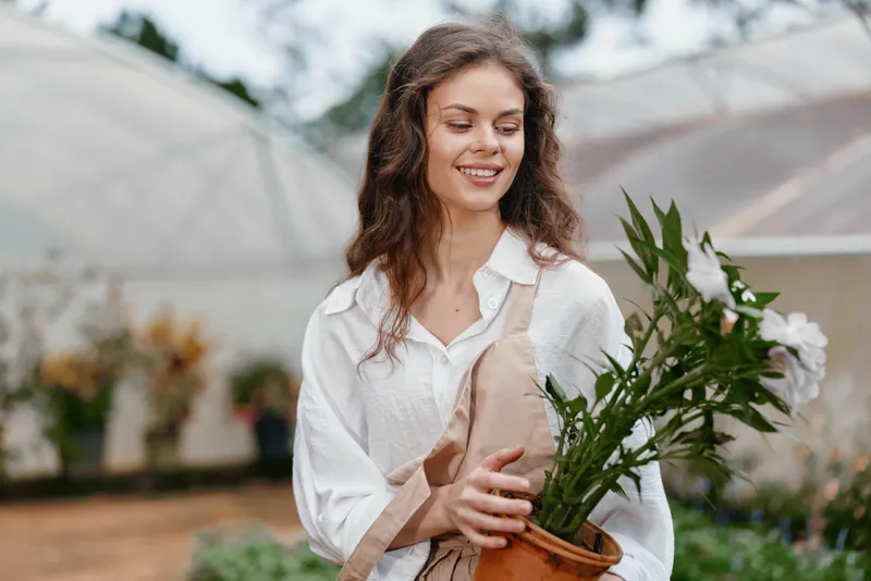 Young woman with brown hair in a white shirt and apron, smiling while holding a potted plant in a greenhouse, symbolizing gardening and nature