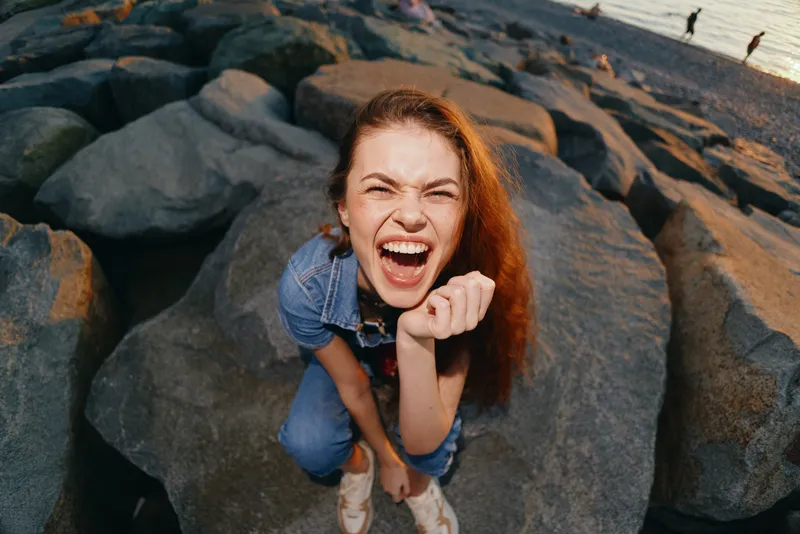 Lifestyle woman in modern boho-western denim street style laughing joyfully with natural red hair and film color tones on rocky beach during sunset.