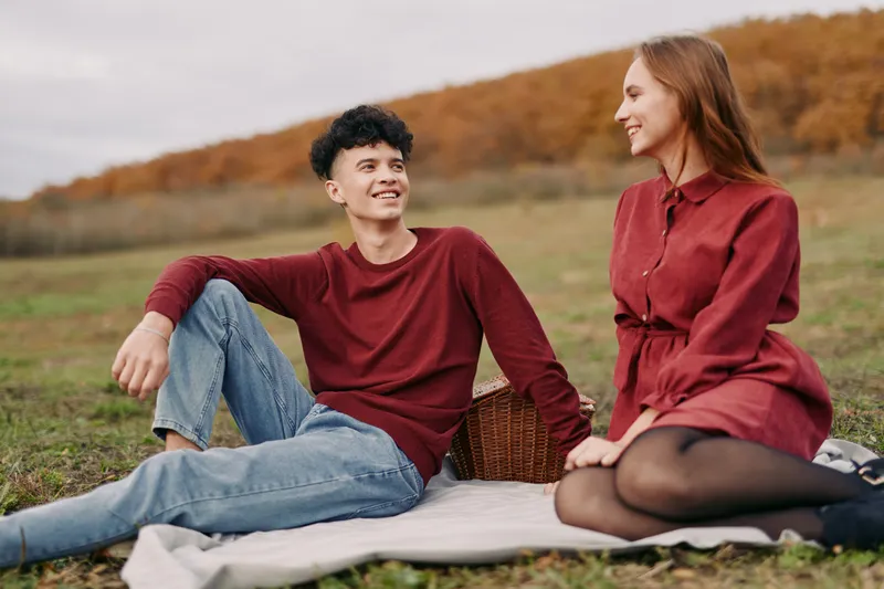 A young couple on a rustic picnic blanket in an autumn field, sharing a genuine, authentic moment of relaxed conversation and warm eye contact, natural expressions and simple joy.