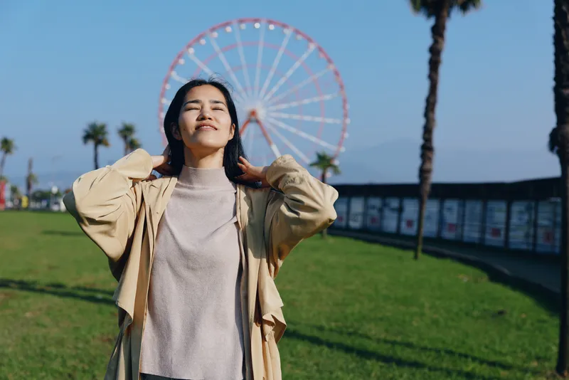 Woman in a light jacket stands outdoors with hands in her hair, a ferris wheel and palm trees in the background, sunny park scenery conveying fashion, leisure and carefree mood, summer vibe.