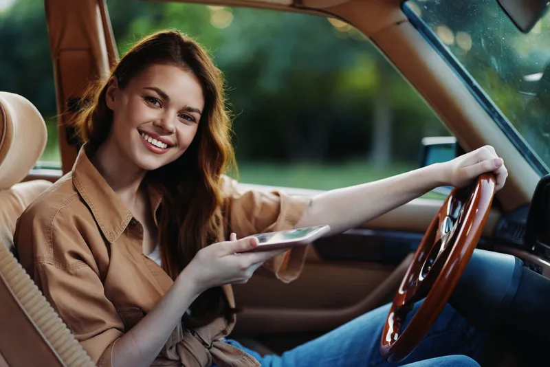 Beautiful young woman smiling inside a car while using a smartphone, dressed casually in a beige shirt, enjoying a sunny outdoor moment.