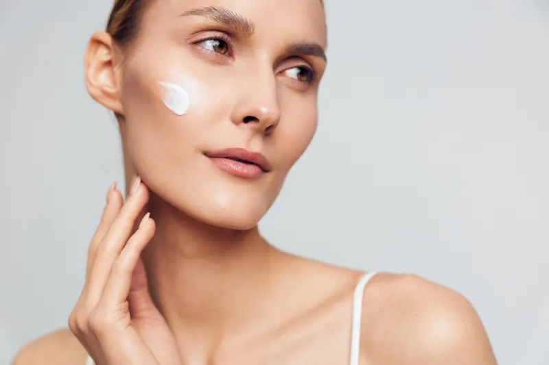 Young woman with a dewy complexion applies cream to her cheek in a studio portrait, with soft lighting highlighting smooth skin and a calm vibe.