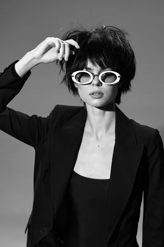 Stylish woman wearing white sunglasses and black blazer posing confidently in black and white portrait