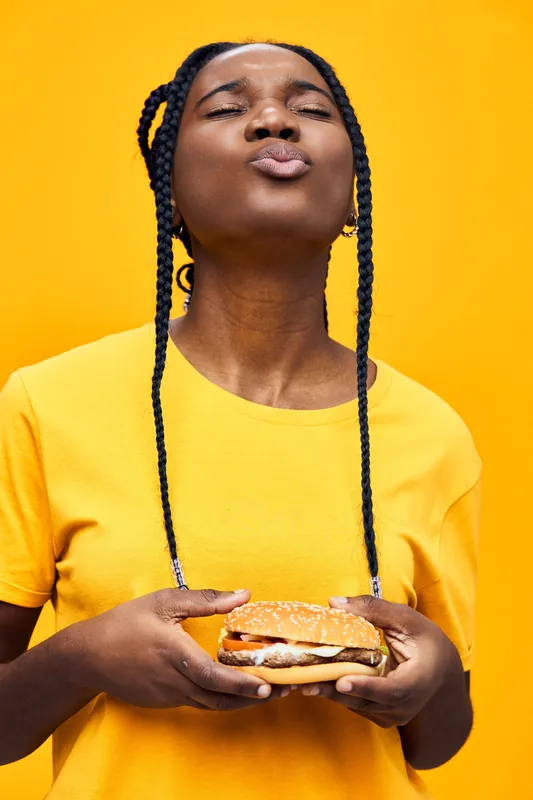 Happy young woman enjoying a delicious burger, showcasing joy in vibrant yellow background, perfect for food and lifestyle concepts