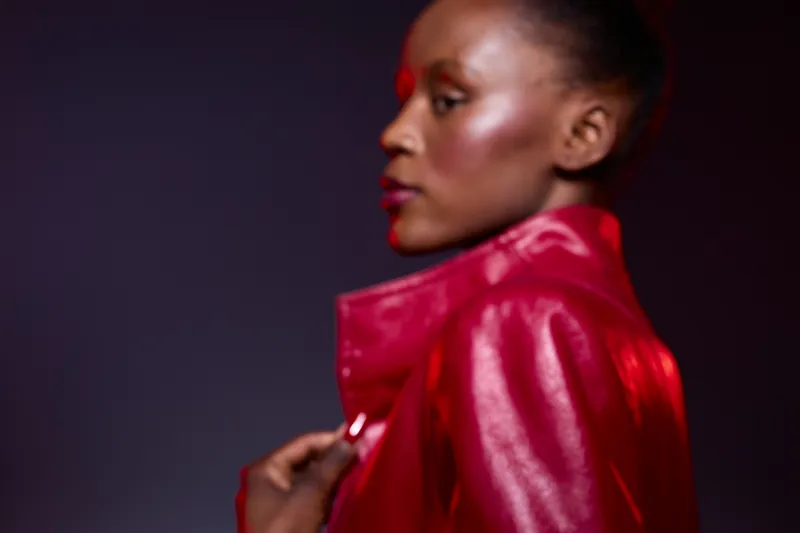 Portrait of a confident black woman with dark skin wearing a shiny red jacket. Side profile on a dark purple background. Concept of fashion, beauty, and elegance in studio lighting.
