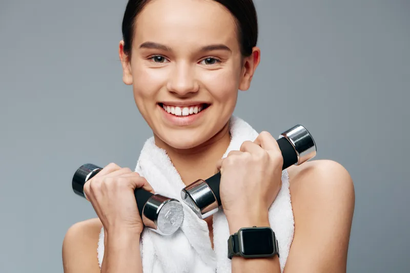 A smiling woman holds dumbbells with a towel draped over her shoulders, ready for a workout. Fitness and sport lifestyle, healthy, athletic energy in a studio setting.