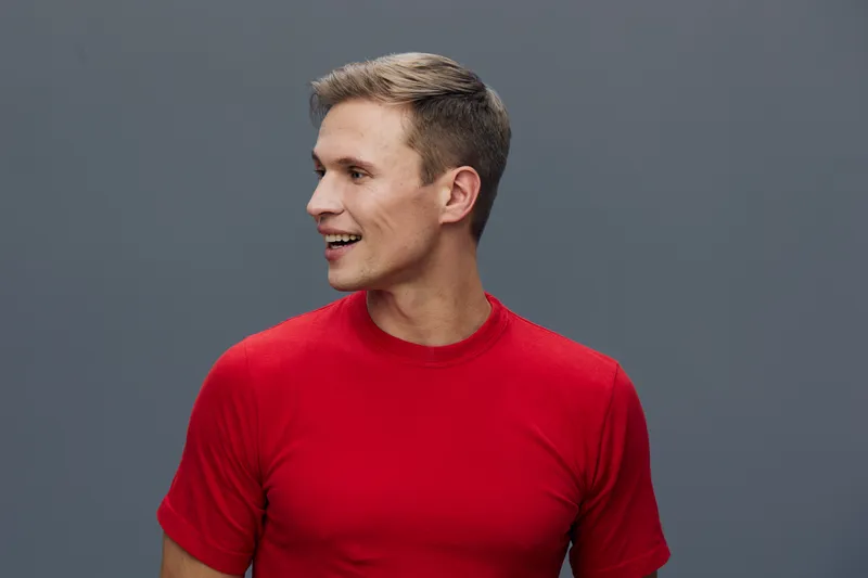 Smiling young man wearing red t-shirt with short hair looking to his side against solid gray background. Studio portrait. Happy, cheerful, casual, fashion, male, confident, studio photo.
