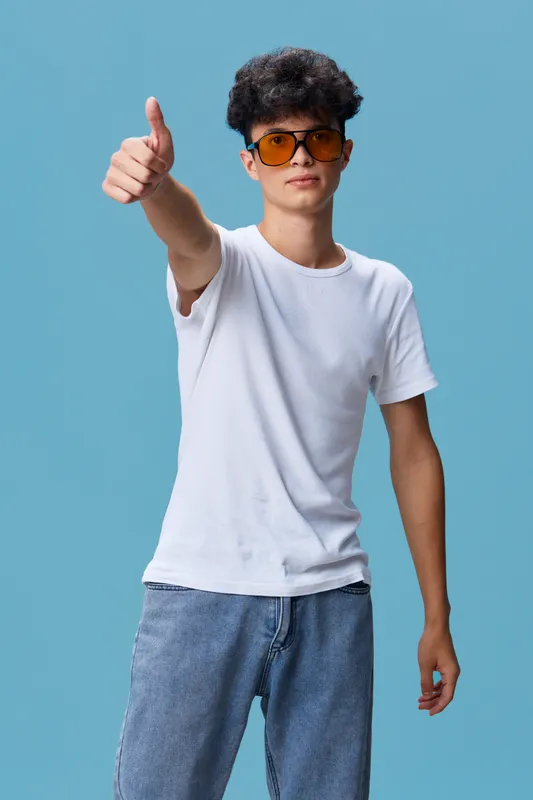 young man in white t-shirt and jeans showing thumbs up gesture with confident expression on blue background, casual style and cool sunglasses