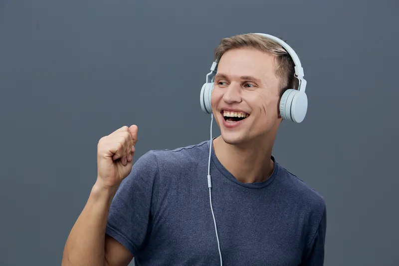 Happy man wearing large white headphones smiling excitedly raising fist in celebration isolated on plain gray background. Lifestyle concept of music enjoyment, joy, entertainment, expression, leisure,