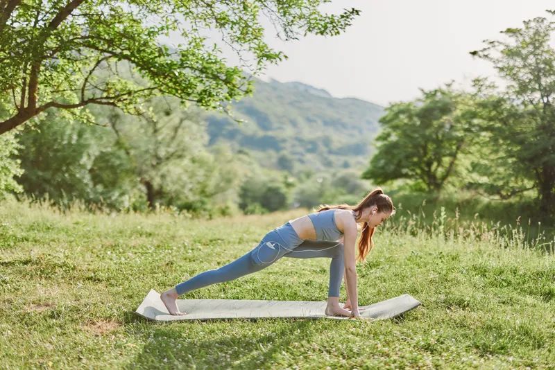 Young woman practicing yoga outdoors on a mat in a serene green landscape, enhancing fitness, mindfulness, and healthy lifestyle