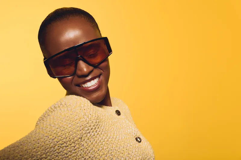 Smiling black woman with dark skin wearing oversized sunglasses and beige knit sweater poses against yellow background. Concept for fashion, confidence, style, and beauty portrait.
