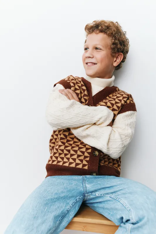 Cheerful young boy sits on a wooden stool, arms crossed, wearing a patterned knit vest and cream sweater, posing against a clean white background with a warm confident smile.