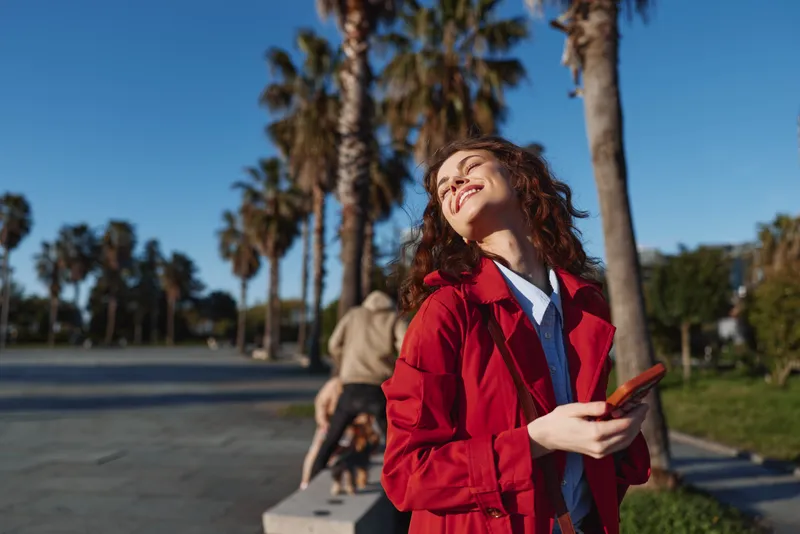 Young woman with curly hair enjoys a sunny day outdoors while holding a smartphone and smiling with closed eyes in a relaxed urban park setting.