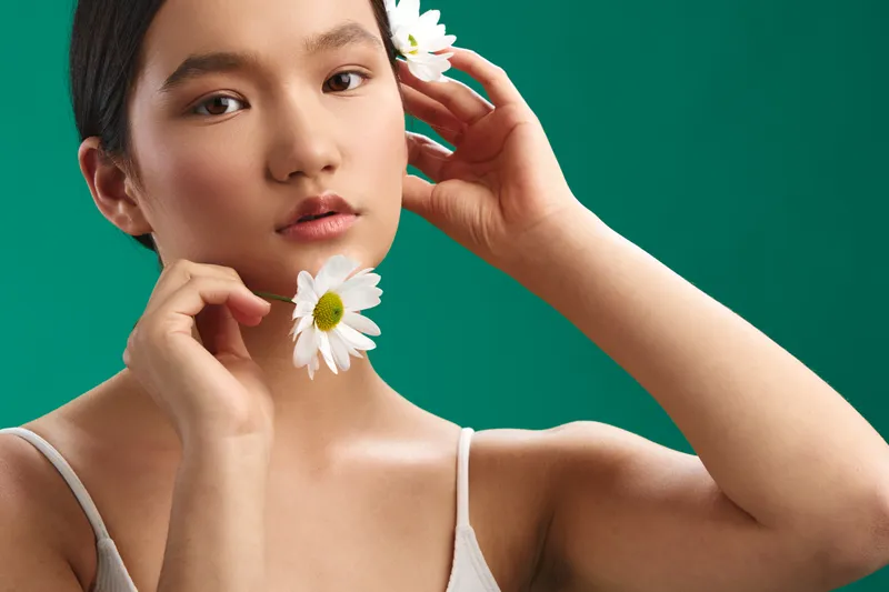 asian woman gazes softly at the camera as she holds white daisies near her face, showcasing natural beauty, calm mood, and studio lighting.