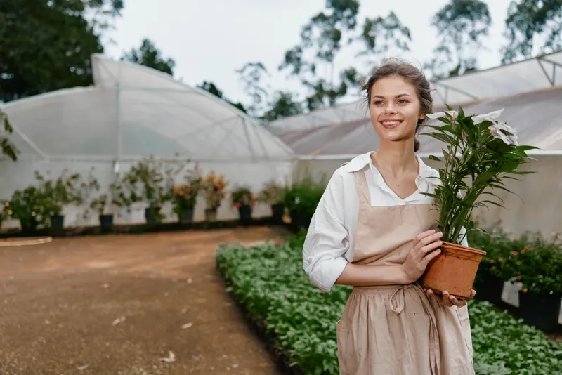 Young female gardener in apron smiling while holding a potted plant in a greenhouse, showcasing passion for gardening and nature