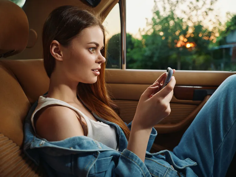 Young woman in casual clothing using a smartphone inside a car during sunset, showcasing a relaxed and joyful expression with a warm color palette.