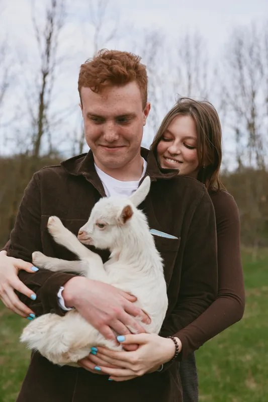 Happy couple holding a fluffy goat kid outdoors in a park, daytime scene. Tender moment of companionship, care, and rural lifestyle. Soft natural light highlights warmth, affection, and bond with