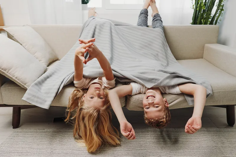Two happy children lie upside down on a sofa covered with a blanket in a bright living room, smiling and playing together. Concept of childhood joy, family time, and cozy home environment.