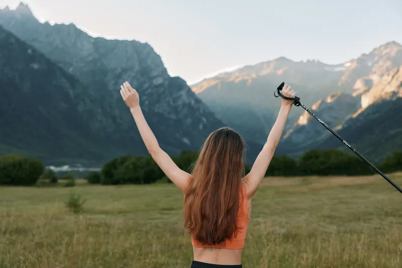 A joyful individual stands in a wide valley surrounded by towering mountains, arms raised in triumph, capturing a moment of achievement and freedom in pristine nature.