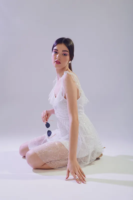 Young woman in a white lace dress poses sitting on the floor holding sunglasses. Studio shot on a light background. Concept for woman beauty fashion and elegance.