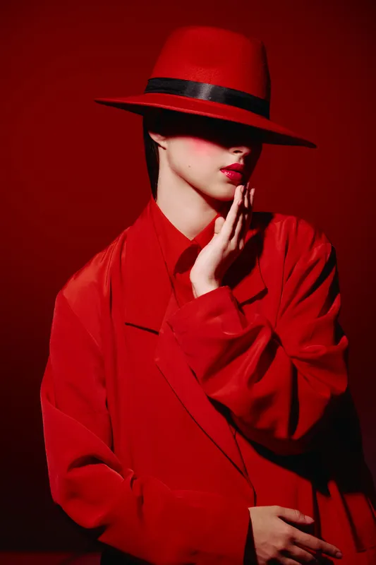 Woman in bright red hat and coat conceals her eyes, highlighting dramatic makeup with red lips and soft lighting. Creative beauty fashion portrait on red background.